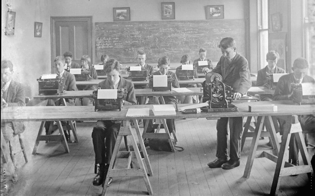 13 boys sitting at tables behind typewriters
