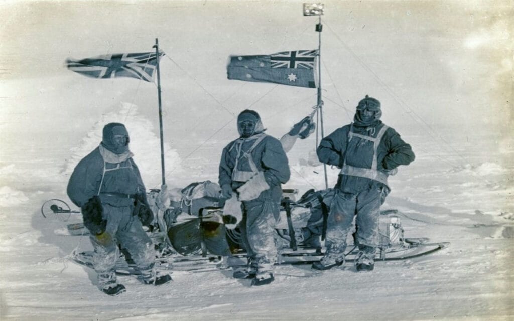 three men in snow clothes, on snowy ground with a sled, an English flag and an Australian flag.