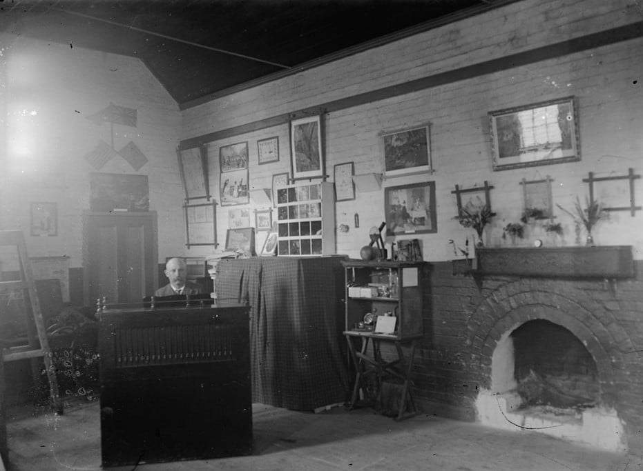 Interior view of hall with brick fireplace, pictures on wall, specimens in display boxes against wall, wooden easel on left; man seated behind an organ or piano. 