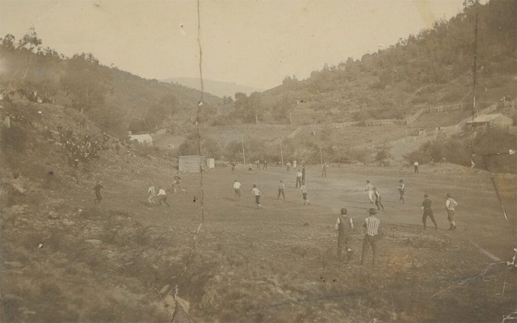 Elevated view of a country football game on a field surrounded by hills, spectators seated on rising ground around the field, fenced off paddocks and buildings in the background.