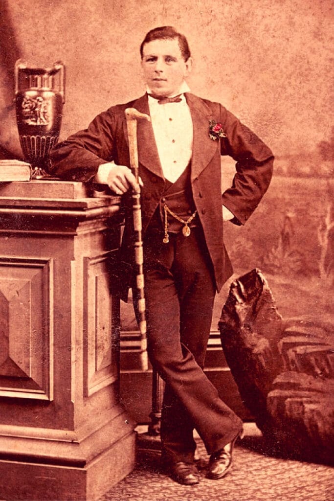 Studio portrait of a young man, full length, full face, standing with right leg crossed in front, left hand on hip, right arm resting on pedestal with hand holding a cane, wearing dinner jacket with vest and bow tie.