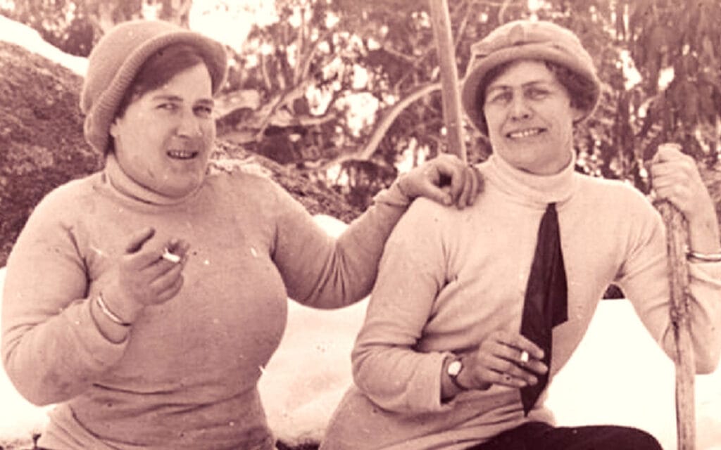 Two women sitting on a fallen log in the snow smoking, one holding a staff.