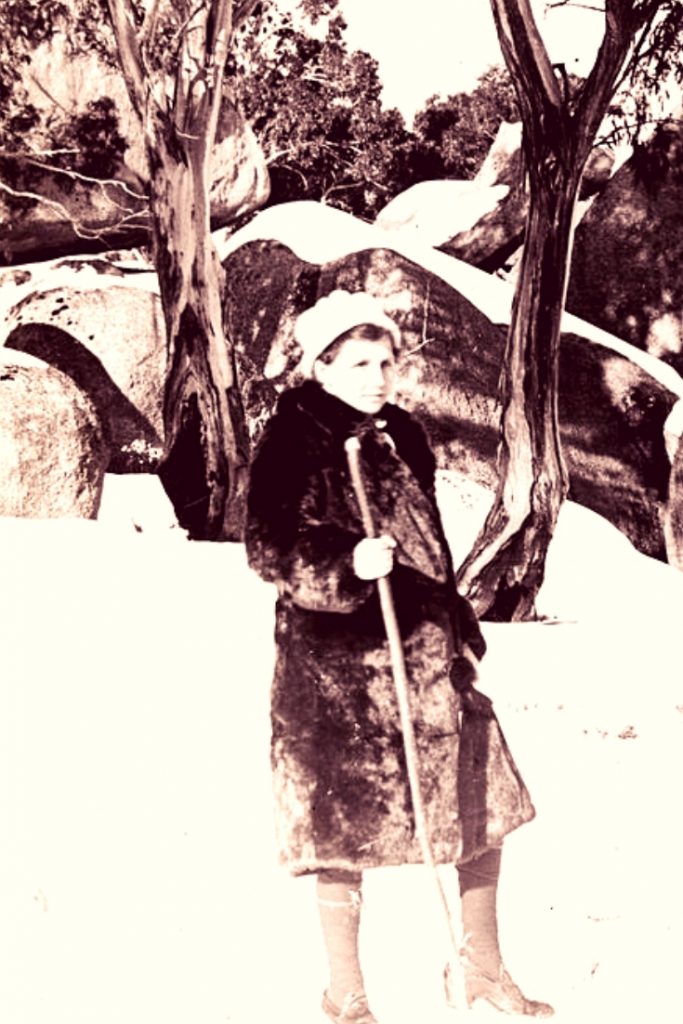 woman in front of snow covered rocks and trees wearing a fur coat, hat and carrying a stick.