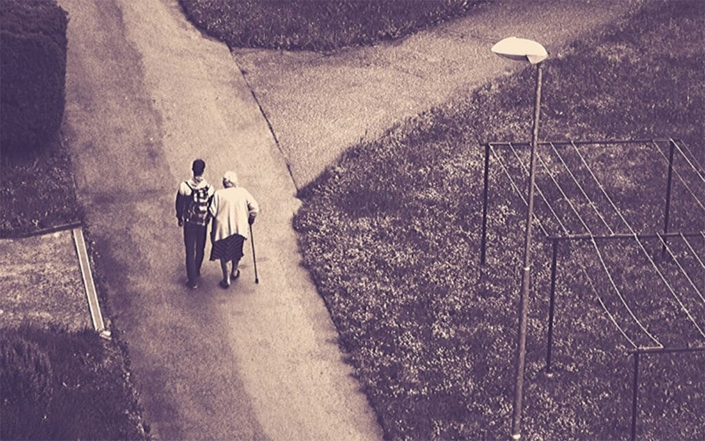 Young man respectfully assisting an older woman with a walking stick as she walks down a path.