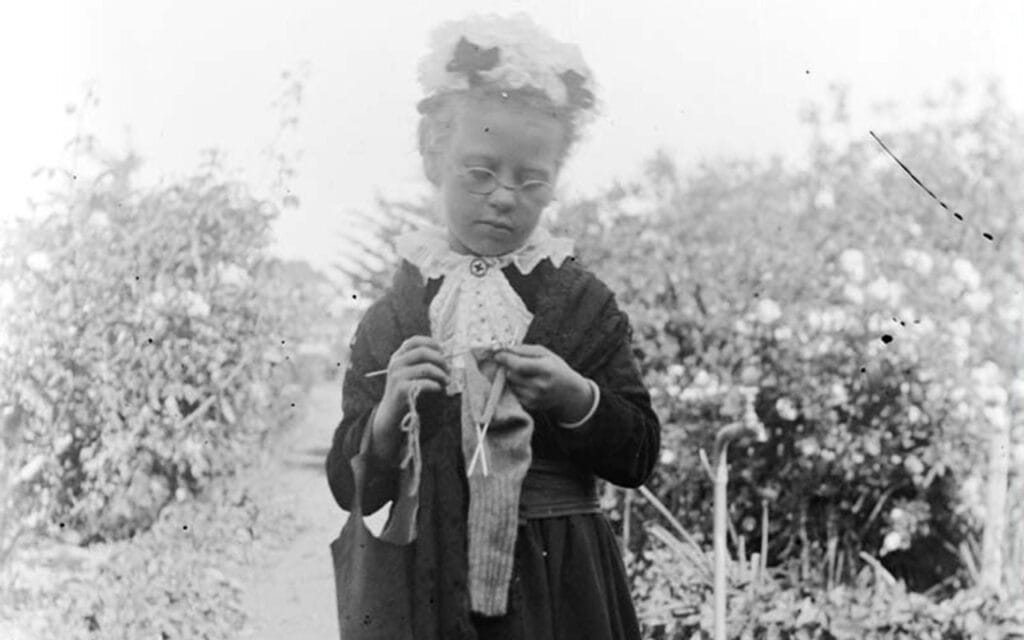 Whole-length portrait of child dressed as elderly woman wearing long dress and hat, knitting as she walks in the garden