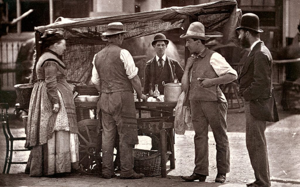 Victorian era photo of a man in a button up shirt with a man in a suit standing before a cart being served by a man and a woman.