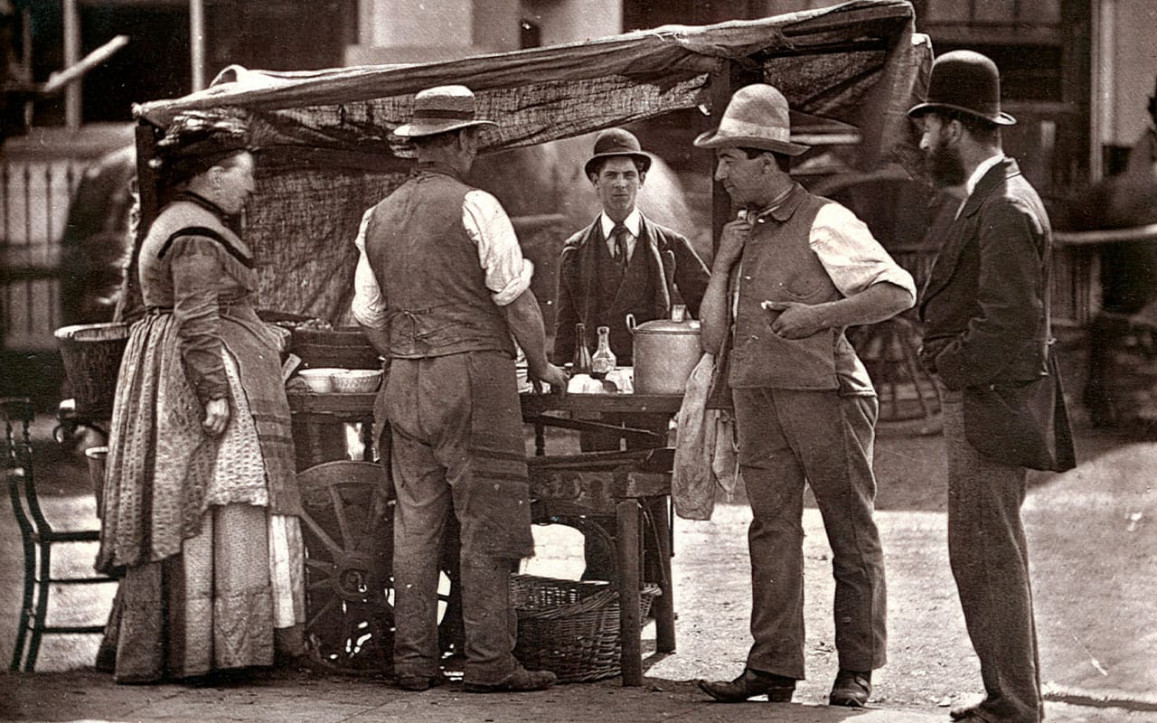 Victorian era photo of a man in a button up shirt with a man in a suit standing before a cart being served by a man and a woman.